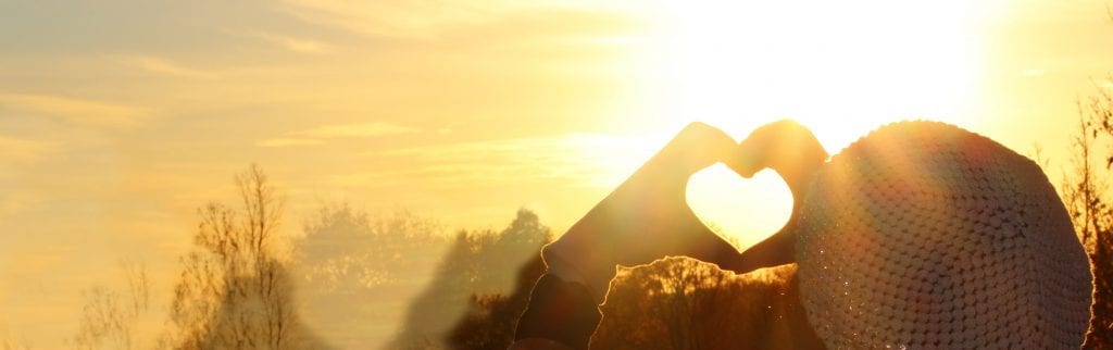 Woman hands in gloves forming a heart symbol, on a beautiful sunset light background symbolising self-love and self-care.