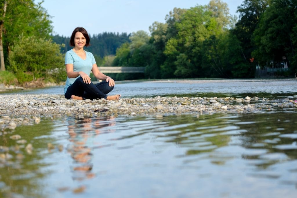Woman sitting by a river in nature, representing calm, connection and holistic wellbeing.