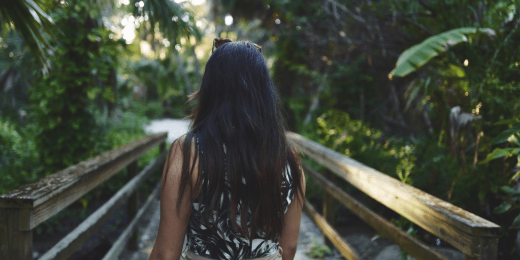 Woman walking confidently over a wooden bridge towards a forest where sun rays shine through.